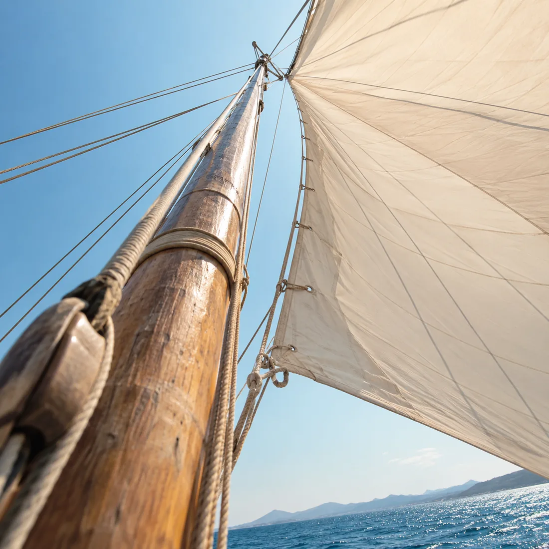 Looking up a wooden mast to a full sail against a clear blue sky, open sea beyond